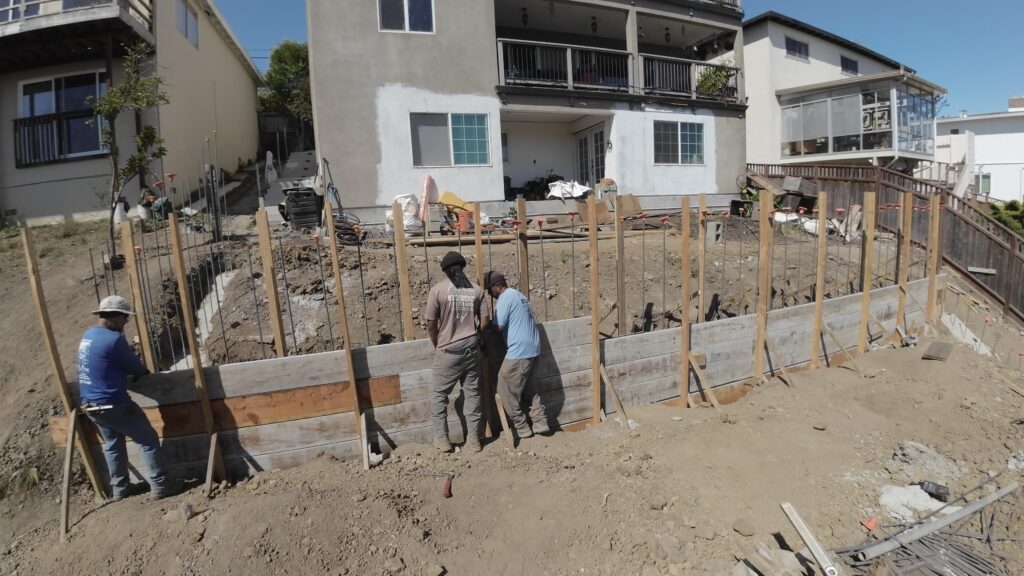 All Access Construction crew forming concrete retaining wall panels in Belmont, showing vertical rebar, wood forms, and hillside layout.