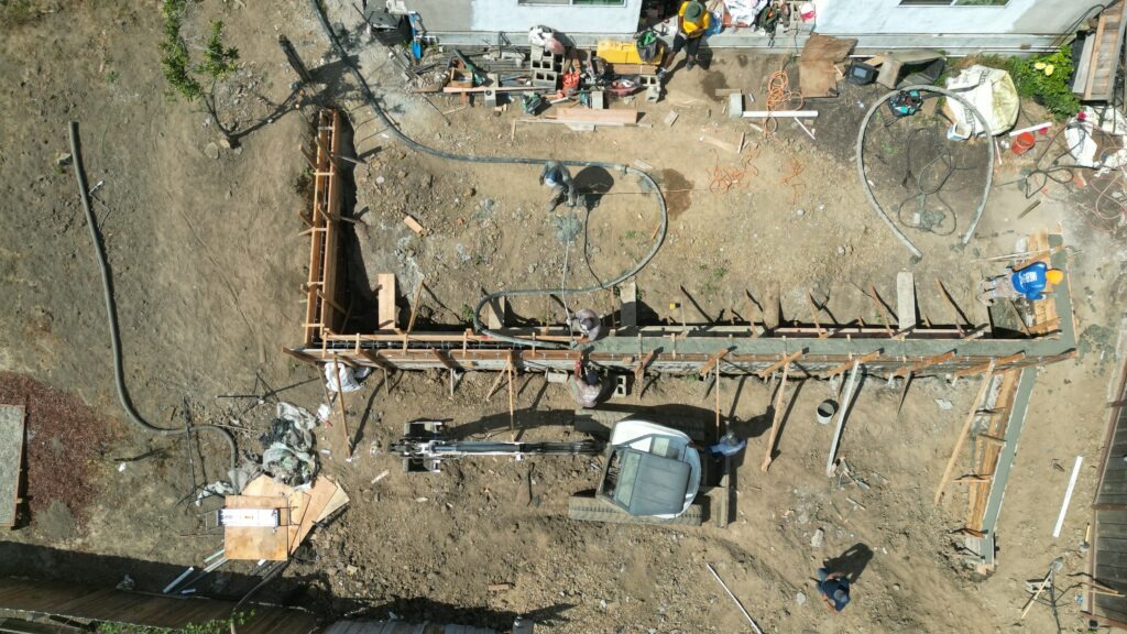 All Access Construction drone shot showing excavator and crew pouring concrete into retaining wall forms on Belmont hillside project.