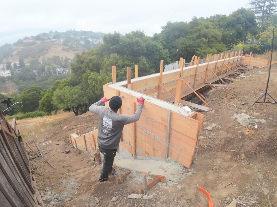All Access Construction removing retaining wall formwork on Belmont hillside project, showing fresh concrete wall and bracing along steep slope.