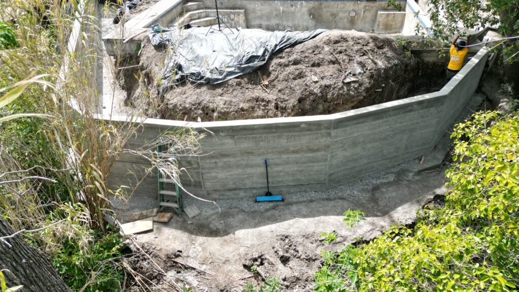 All Access Construction concrete retaining wall in Walnut Creek, Contra Costa County—freshly stripped forms, broom-clean base, stepped hillside excavation, and worker setting alignment line for final backfill prep