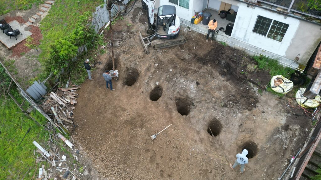 All Access Construction drilling 20-foot deep pier holes on a steep Belmont hillside using a Bobcat excavator during the retaining wall foundation phase.