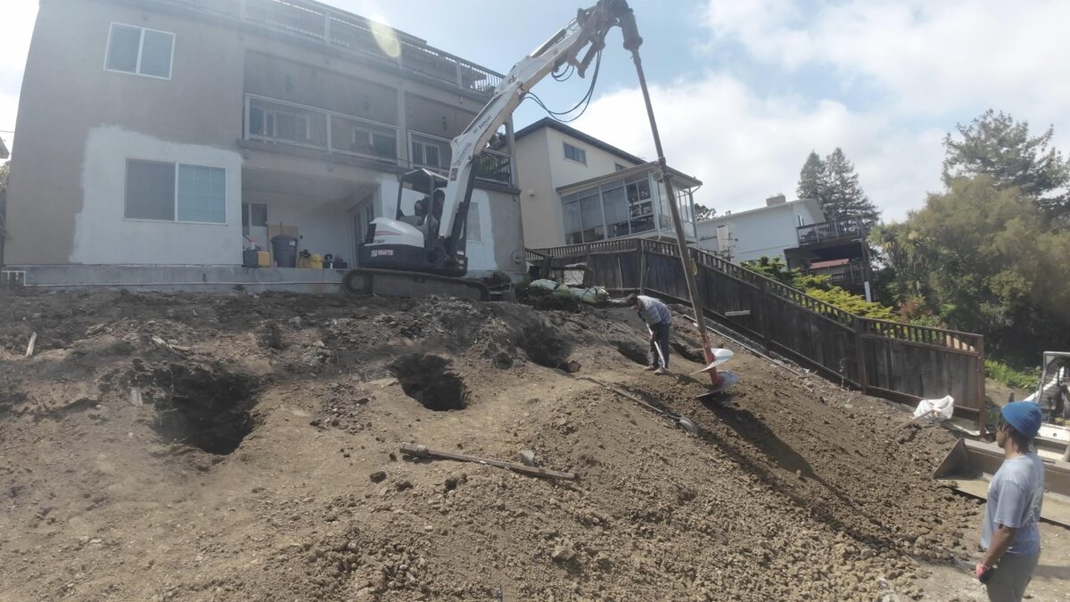 All Access crew drilling deep concrete piers on a Belmont hillside slope using an extended auger rig for the structural retaining wall system.