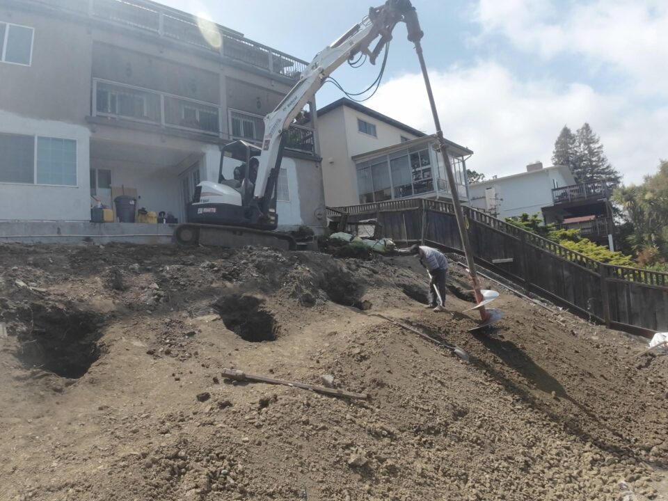 All Access crew drilling deep concrete piers on a Belmont hillside slope using an extended auger rig for the structural retaining wall system.